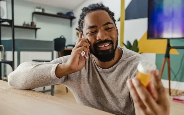 Smiling man holding a pill bottle while talking on the phone in a cozy room.